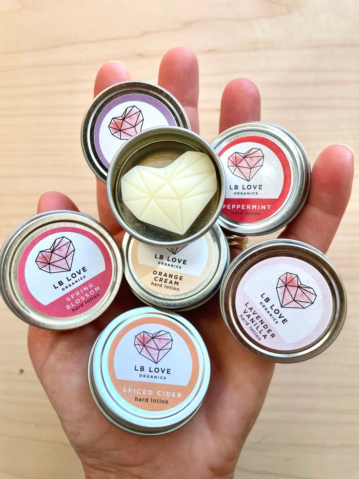 Hand holding multiple tins of LB Love Organics hard lotion with visible cream inside.