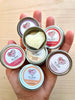 Hand holding multiple tins of LB Love Organics moisturizing hard lotion with visible heart shaped lotion 